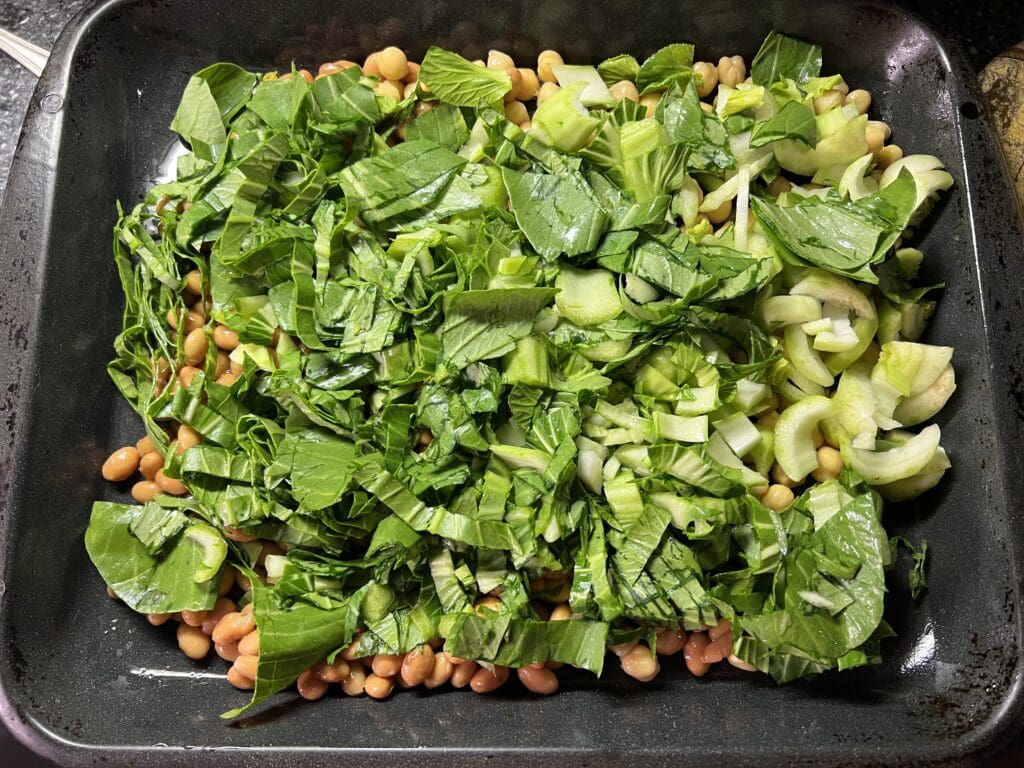 Sliced bok choy, celery, leafy greens spread over beans in a roasting pan.