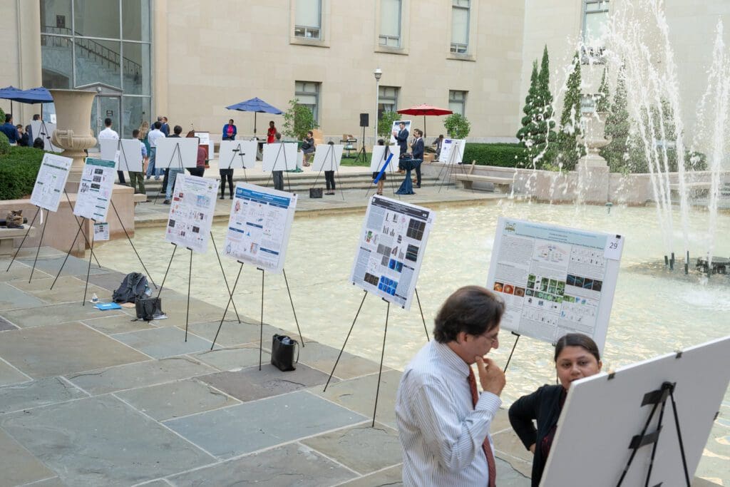 Multiple science poster displays in front of fountain. People perusing posters.