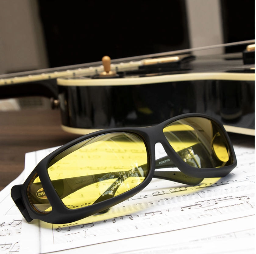 Cocoons wear-over yellow filter glasses for macular degeneration sit on top of a music sheet with a guitar in the background. 