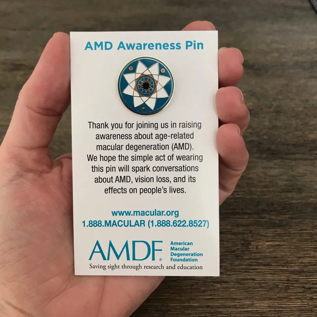 Hand holding card with AMD Awareness Pin attached. Text reads: Thank you for joining us in raising awareness about age-related macular degeneration (AMD). We hope the simple act of wearing this pin will spark conversations about AMD, vision loss, and its effects on people's lives.